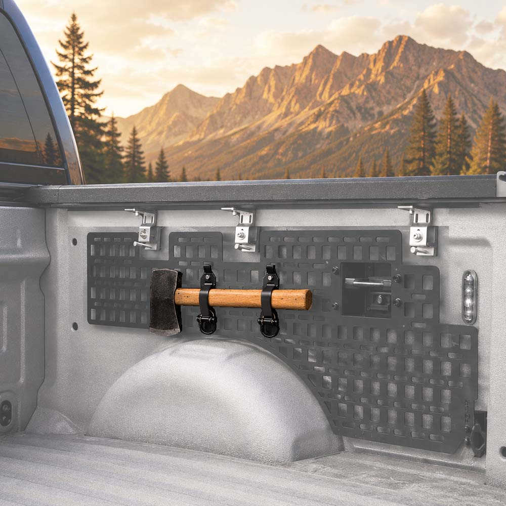 A set of black textured truck bed racks with silver mounting clips installed in the bed of a pickup truck, with a wooden handlebar and a hammer hanging on the rack. The background shows a scenic mountain landscape at sunset.