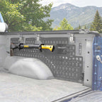 A black and yellow DOWNS firearm rack mounted on the tailgate of a pickup truck, shown outdoors with mountains and trees in the background.