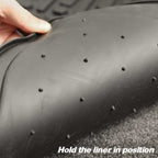 Close-up of a black rubber vehicle liner with small raised nubs, being held by a hand to show flexibility, with the text 'Hold the liner in position' at the bottom.