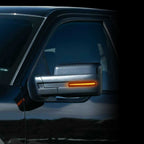 Close-up of a vehicle side mirror with an illuminated amber side marker light, mounted on a dark car body.