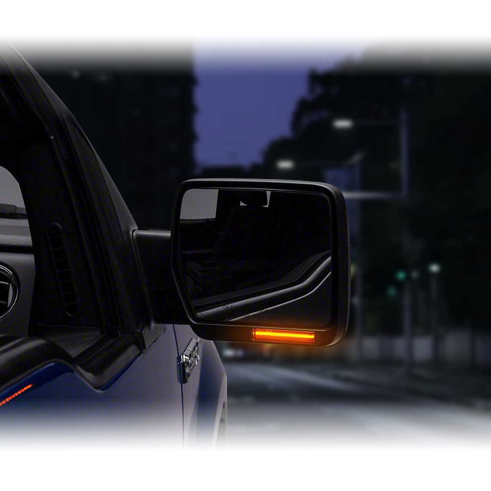 A close-up of a vehicle side mirror with an illuminated amber side marker light, attached to a dark blue truck at night on a blurred road scene.