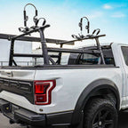 A black dual-bike carrier rack mounted on the bed of a white Ford Super Duty pickup truck, shown from the rear three-quarter angle against a blue sky and outdoor background.
