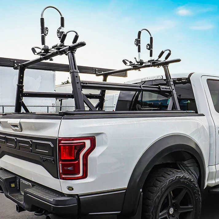 A black dual-bike carrier rack mounted on the bed of a white Ford Super Duty pickup truck, shown from the rear three-quarter angle against a blue sky and outdoor background.