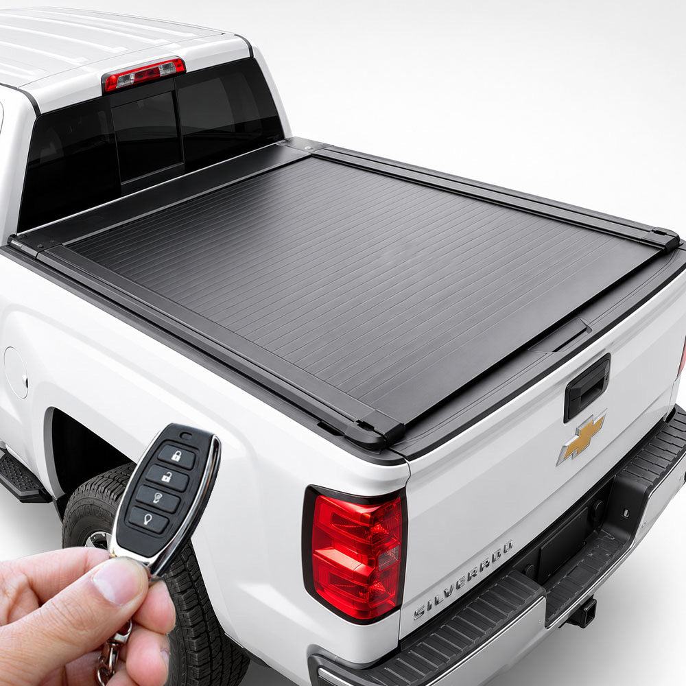 A black retractable truck bed cover rolled up over the bed of a white pickup truck, with a hand holding a key fob in the foreground on a clean white studio background.