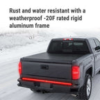 A dark gray pickup truck driving on a snowy road, shown from the rear three-quarter angle. The bed is covered with a black tonneau-style cover, and a red LED light bar is illuminated along the lower rear bumper.