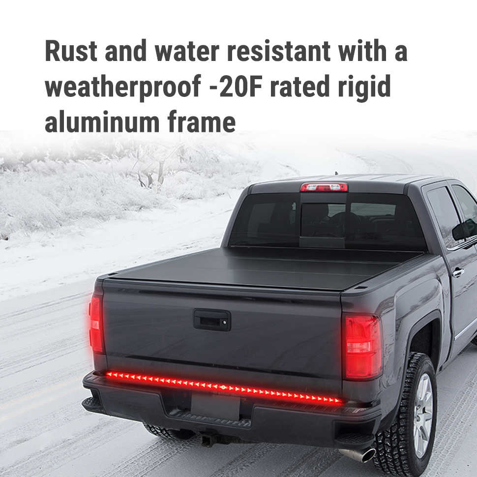 A dark gray pickup truck driving on a snowy road, shown from the rear three-quarter angle. The bed is covered with a black tonneau-style cover, and a red LED light bar is illuminated along the lower rear bumper.