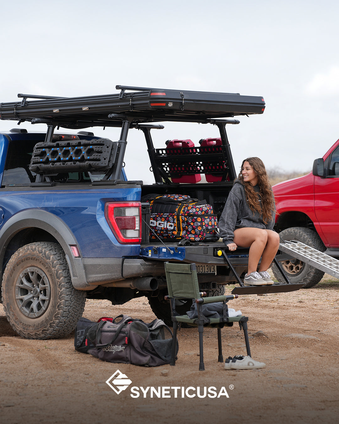 Person sitting on a truck bed with outdoor gear and SYNETICUSA logo.
