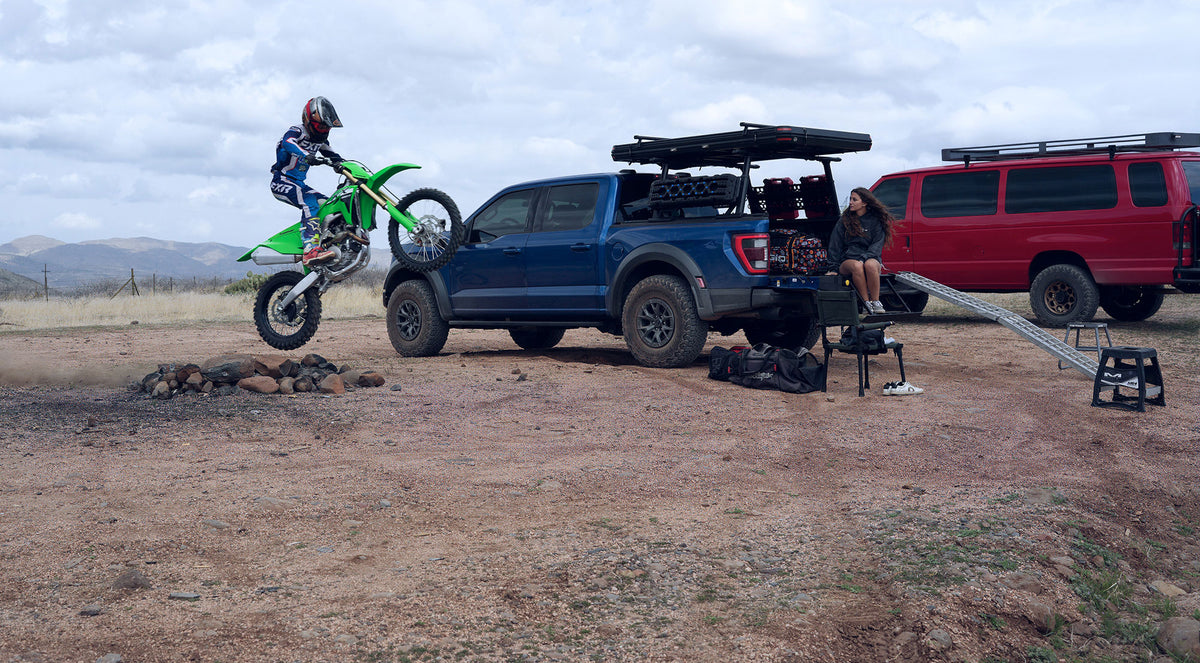 Person on a dirt bike next to a blue truck and red van in an open area with mountains in the background.