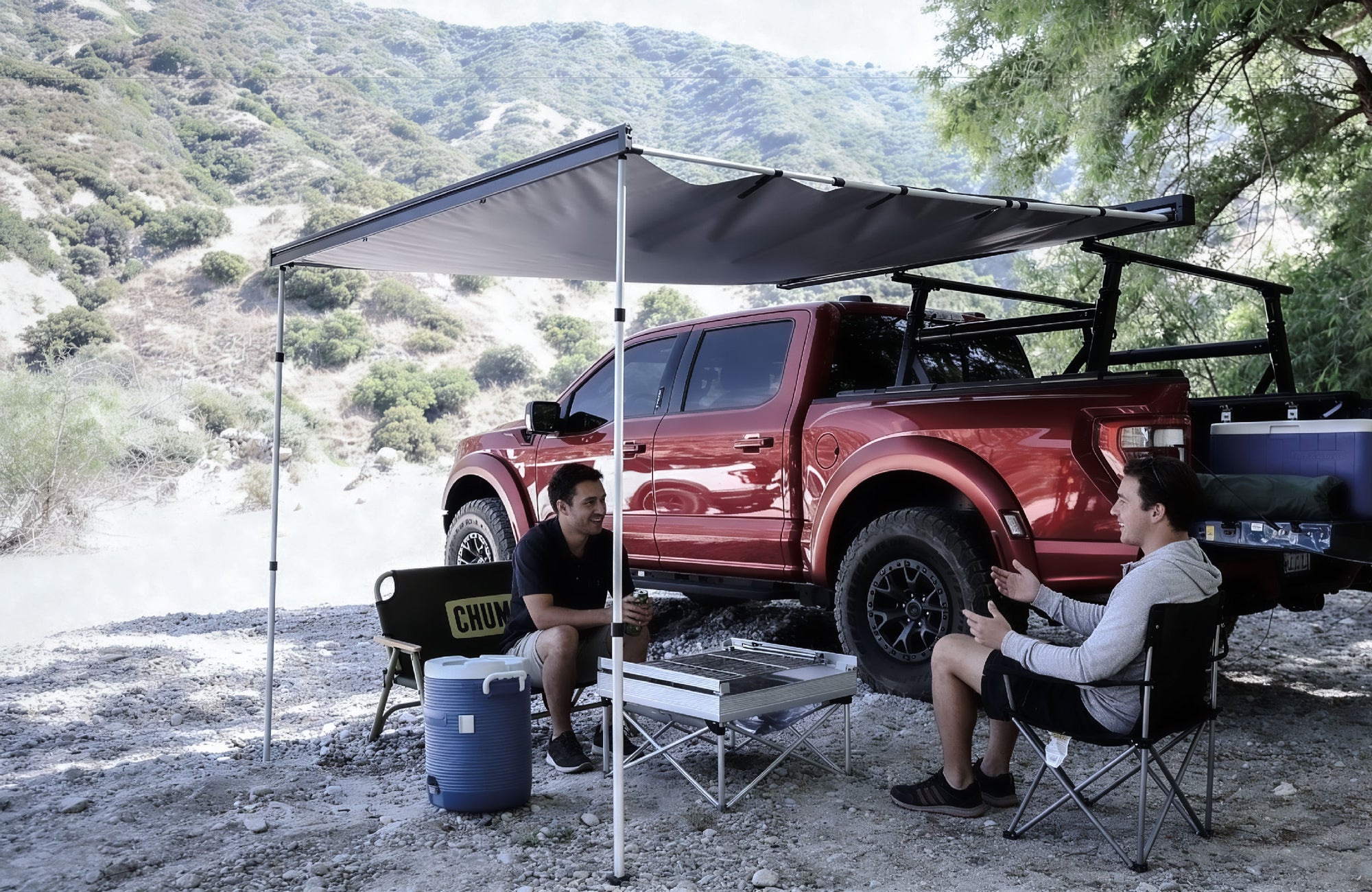 Two people sitting under a portable canopy next to a red pickup truck in a natural setting.